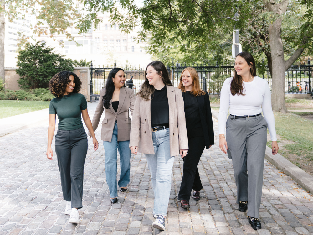 Five women from a Toronto PR and marketing agency walking outdoors on a tree-lined cobblestone path, smiling and talking.